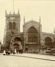 Sepia photo of a large stone church with a clock tower, seen from a cobbled street with people walking in front